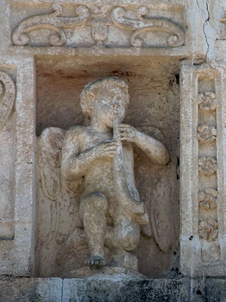 Façade niche sculpture, musician youth (left) - Façade, exterior buttressing, lateral portal, open & posa chapels, atrio & cross