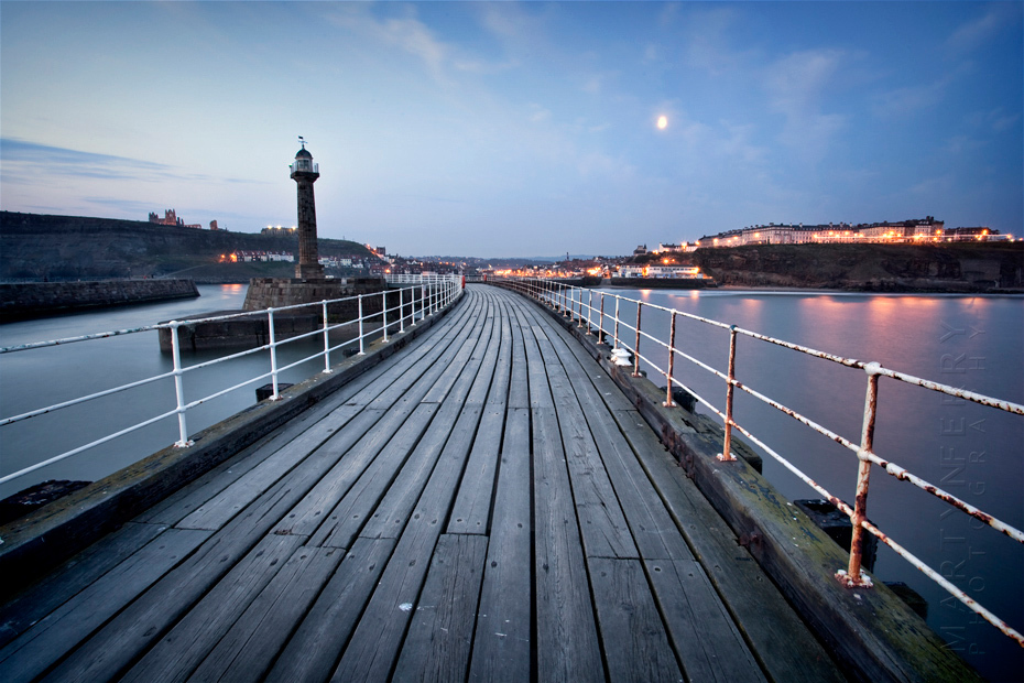 A view along Whitby pier to the lights of the town under an early morning moon
