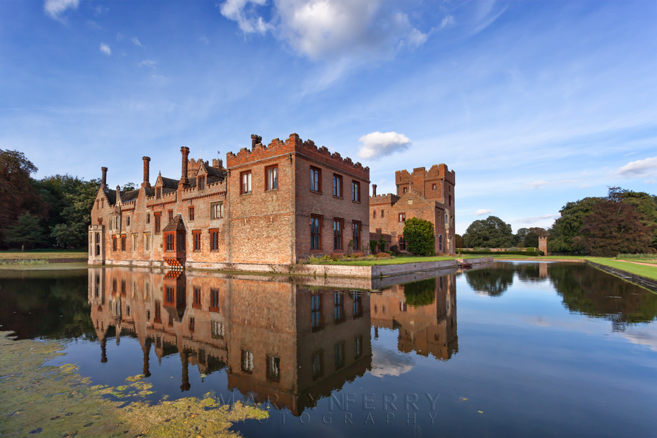 Oxburgh Hall bathed in evening sunlight is reflected in the moat